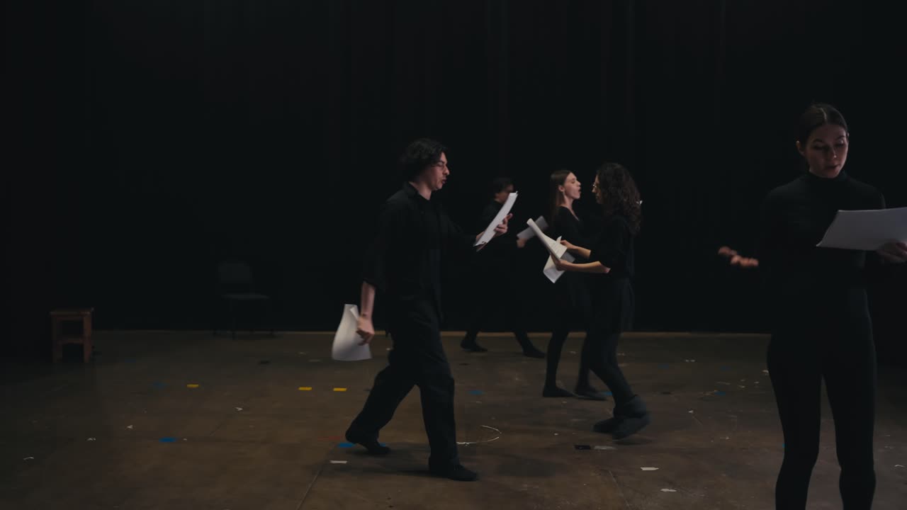 A group of confident actors in black suits walk around the stage and read their words from sheets of paper during their rehearsal and preparation for a performance on stage in a large theater with black curtains closed