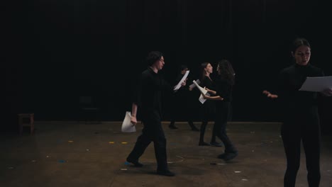 a group of confident actors in black suits walk around the stage and read their words from sheets of paper during their rehearsal and preparation for a performance on stage in a large theater with black curtains closed