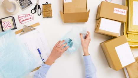 Frau-Packt-Tasse-In-Paketkasten-Bei-Der-Post-9.Lieferung,-Postdienst,-Personen--Und-Versandkonzept-Frau-Verpackt-Tasse-In-Schützende-Luftpolsterfolie-Und-Packt-Sie-In-Paketkasten,-Dann-Streicht-Sie-Den-Artikel-Auf-Dem-Klemmbrett-Bei-Der-Post-Durch