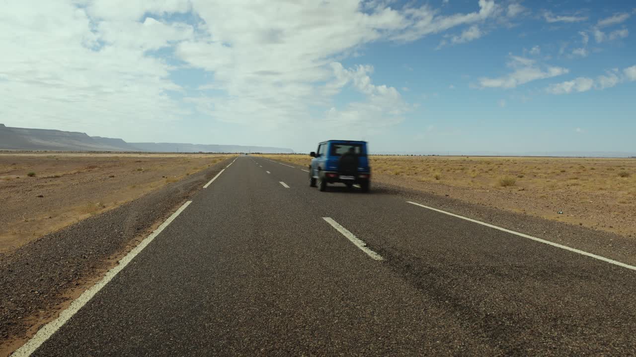 Recta carretera desértica que se extiende hacia el horizonte con un vehículo solitario conduciendo a través de Marruecos, un coche todoterreno azul pasa zumbando.
