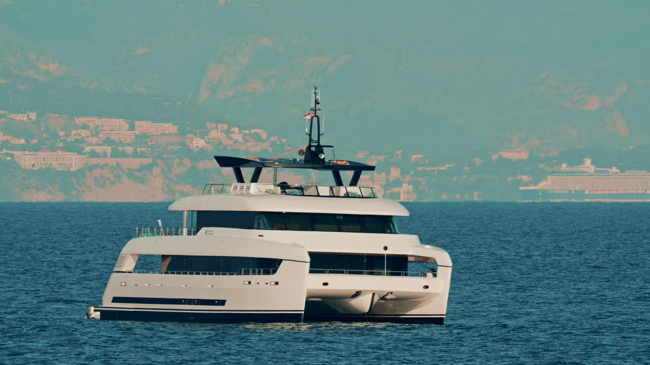 Un moderno yacht catamarano bianco ancorato su acque cristalline con una città costiera sullo sfondo