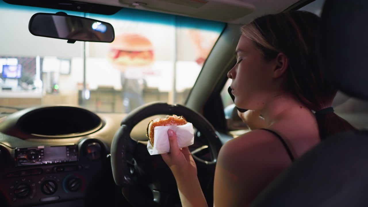 Woman in black top eating burger while in parked car at night, looking contemplative. Interior lights softly illuminating her face, fast-food restaurant visible in background through the windshield