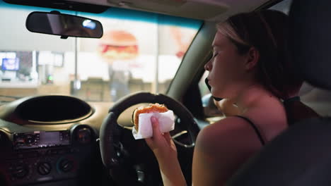 woman in black top eating burger while in parked car at night, looking contemplative. interior lights softly illuminating her face, fast-food restaurant visible in background through the windshield