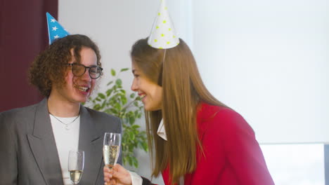 woman with champagne glass talking and laughing with two colleagues while man approaching and offering her a birthday cake at the office