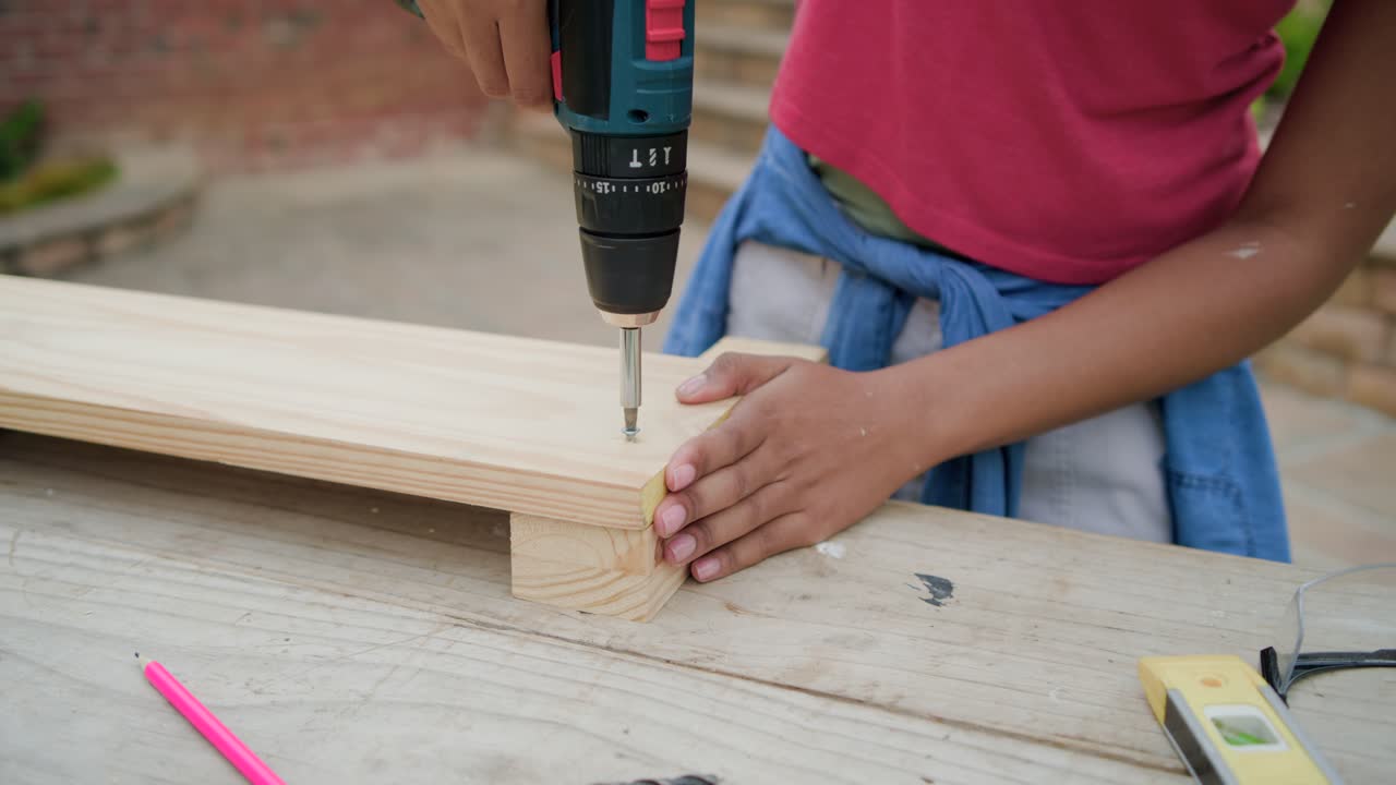 Donna africana con una maglietta rossa che allinea assi, inserisce una vite usando un trapano, costruendo una piattaforma su una panca da patio.