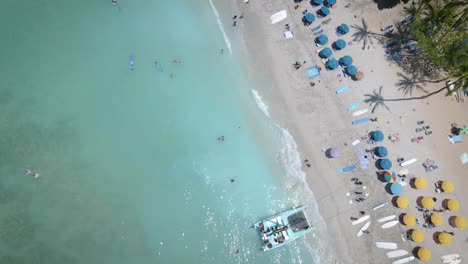 A-drone-captures-the-idyllic-scene-of-a-white-beach,-vibrant-umbrellas,-and-people-swimming-in-the-clear,-inviting-sea