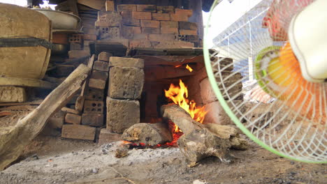 Low-wide-view-of-raging-wood-fire-in-pottery-kiln,-fan-blows-fire,-Than-Ha-Hoi-An-Vietnam