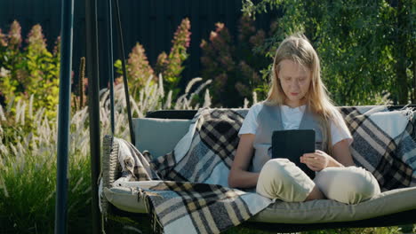 a teenage girl uses a tablet, sits on a garden swing in the backyard of a house.