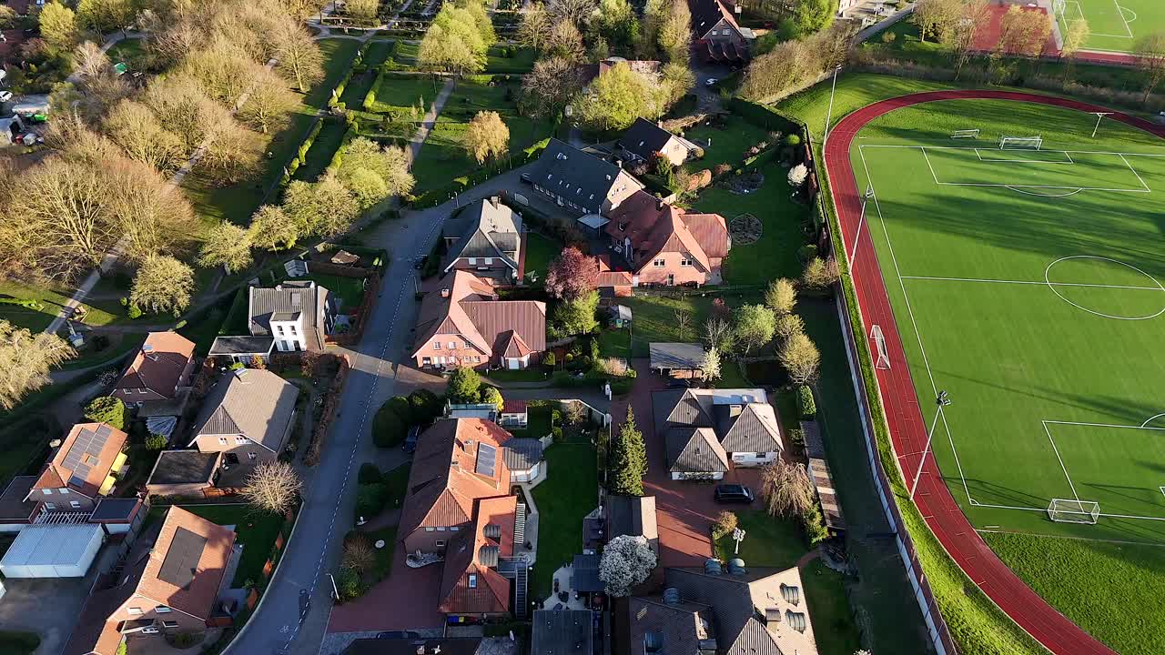 Housing area with solar panels on roof beside soccer field at sunset.Aerial top down flyover shot. Golden sunset with graveyard cemetery in distance.