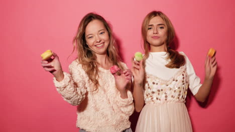 two women holding macaroons in front of a pink background