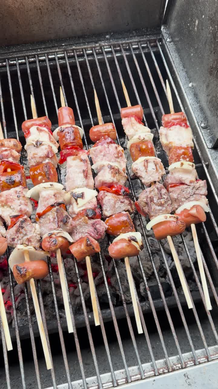 Vertical video panning left to right of meat skewers on a charcoal grill, with red peppers, white onions, and sausages. Glowing red coals and light smoke create a rustic, savory scene.