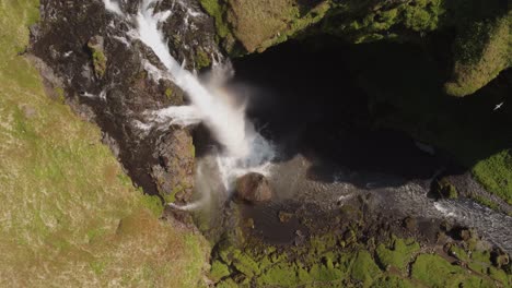 Toma-Aérea-De-Arriba-Hacia-Abajo-De-Una-Hermosa-Cascada-En-Islandia-En-Un-Día-Soleado-Con-Pájaros-Volando-Y-Un-Pequeño-Arco-Iris-Frente-A-Los-Acantilados-Y-Rocas-Verdes-Cubiertos-De-Musgo