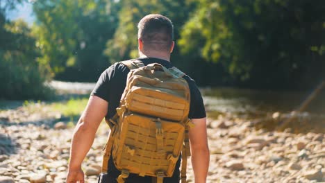 Young-male-traveler-walking-beside-stream-in-forest-walking-on-stones-surounded-by-trees-hike-in-jungle-wild-forest-hot-summer-day,-people-and-wild-nature-in-slow-motion