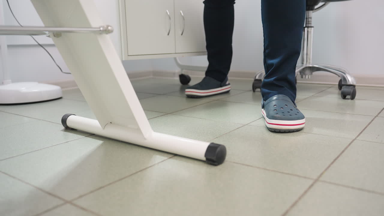 Leg view of aesthetician in crocs and navy blue uniform standing beside clinical bed under bright lamp with chair and product table visible in clean tiled treatment room with stainless trolley