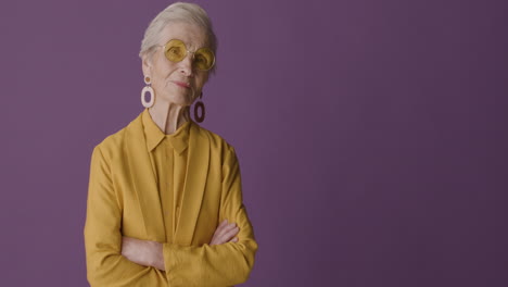 senior woman with short hair wearing mustard colored shirt and jacket, earrings and sunglasses posing with crossed arms and looking at camera on purple background