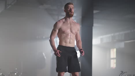 slow motion: muscular shirtless fit man energetically box jumps in hardcore gym doing part of his cross fitness training plan. man is sweaty from intense workout