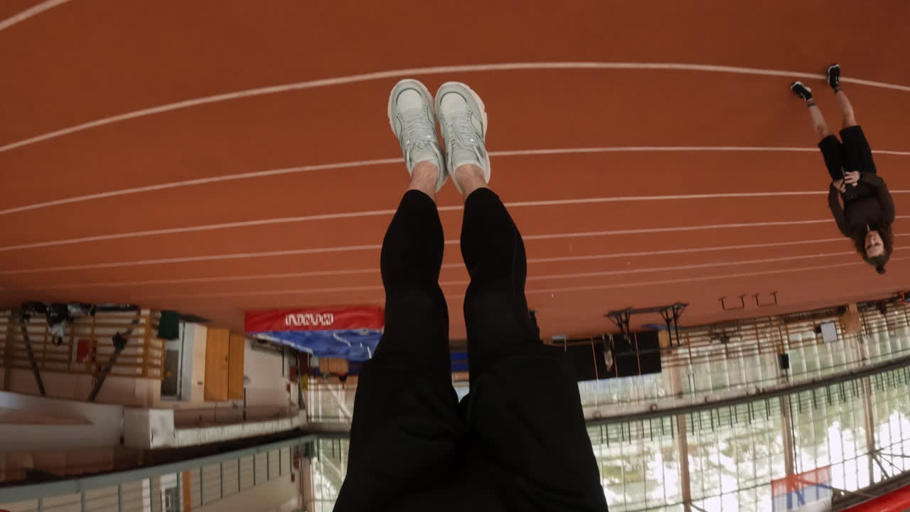 Person performing hanging exercises and pull-ups on a red bar over an indoor running track, often from an inverted perspective