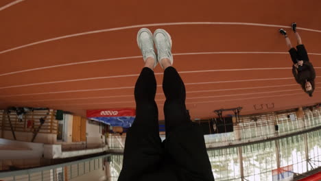 person performing hanging exercises and pull-ups on a red bar over an indoor running track, often from an inverted perspective