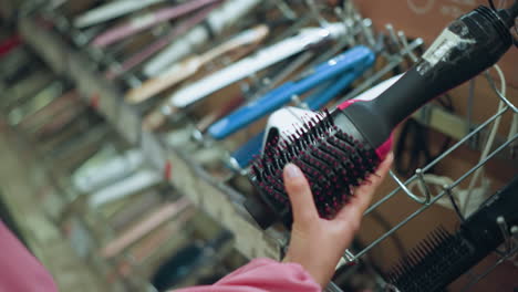 hand of a female shopper closely examining a hair styling tool in a salon store, the shopper is holding a brush-like hair device, observing its details, with a rack of other hair tools