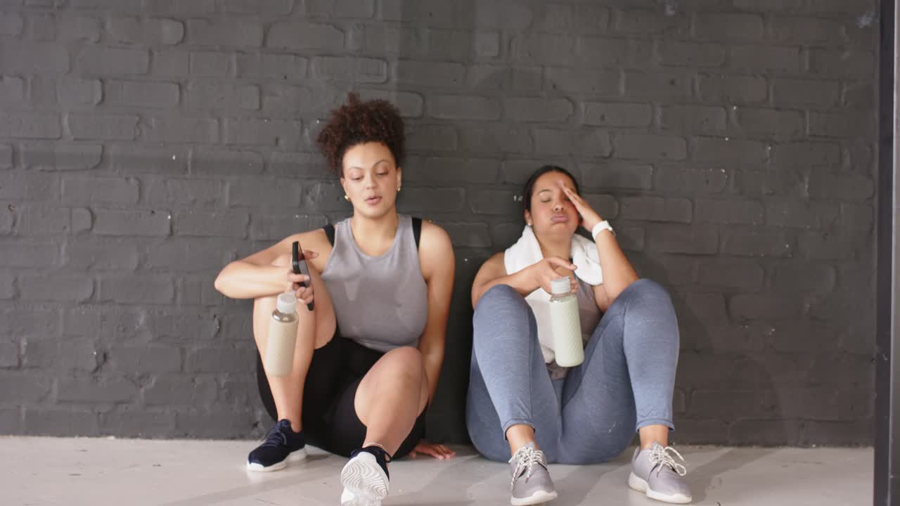 Selesai melakukan squat, rekan latihan wanita meluncur ke bawah sambil minum dari botol air dan mengobrol di gym