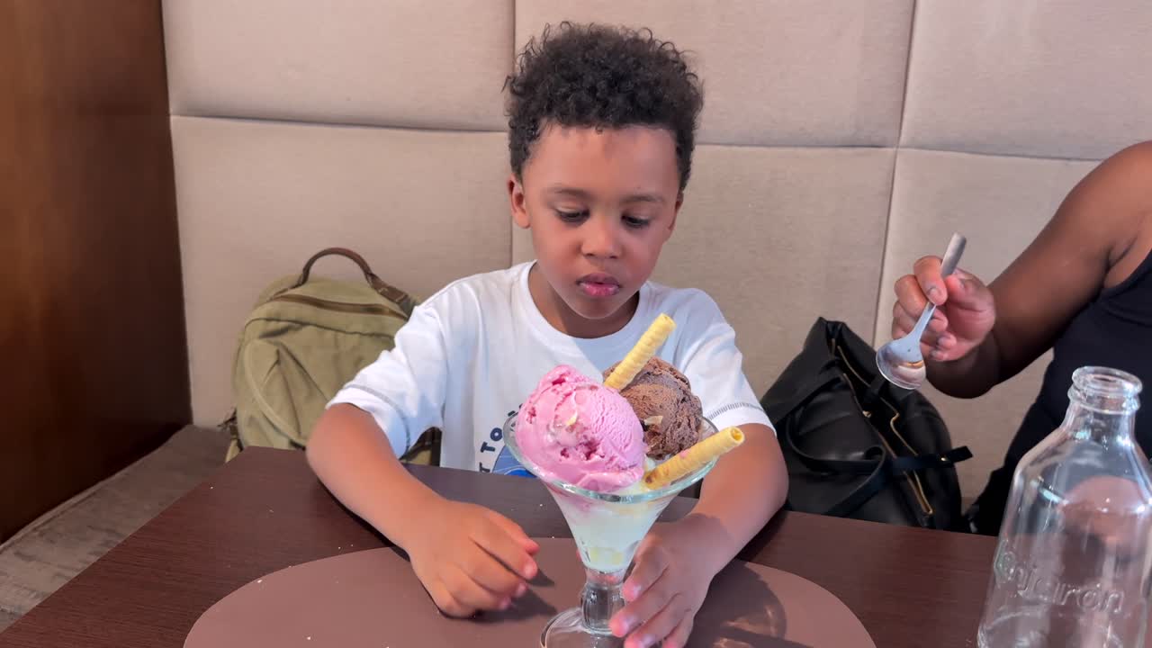 medfium-close up view of a biracial black child sharing with his mother sitting next to him a large ice cream sundae with three scoops of flavors inddor. handheld camera