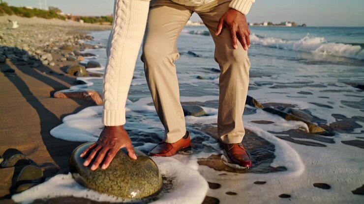 Uomo che cammina con un maglione color crema su una costa rocciosa con la bassa marea, cercando l'equilibrio e toccando un masso