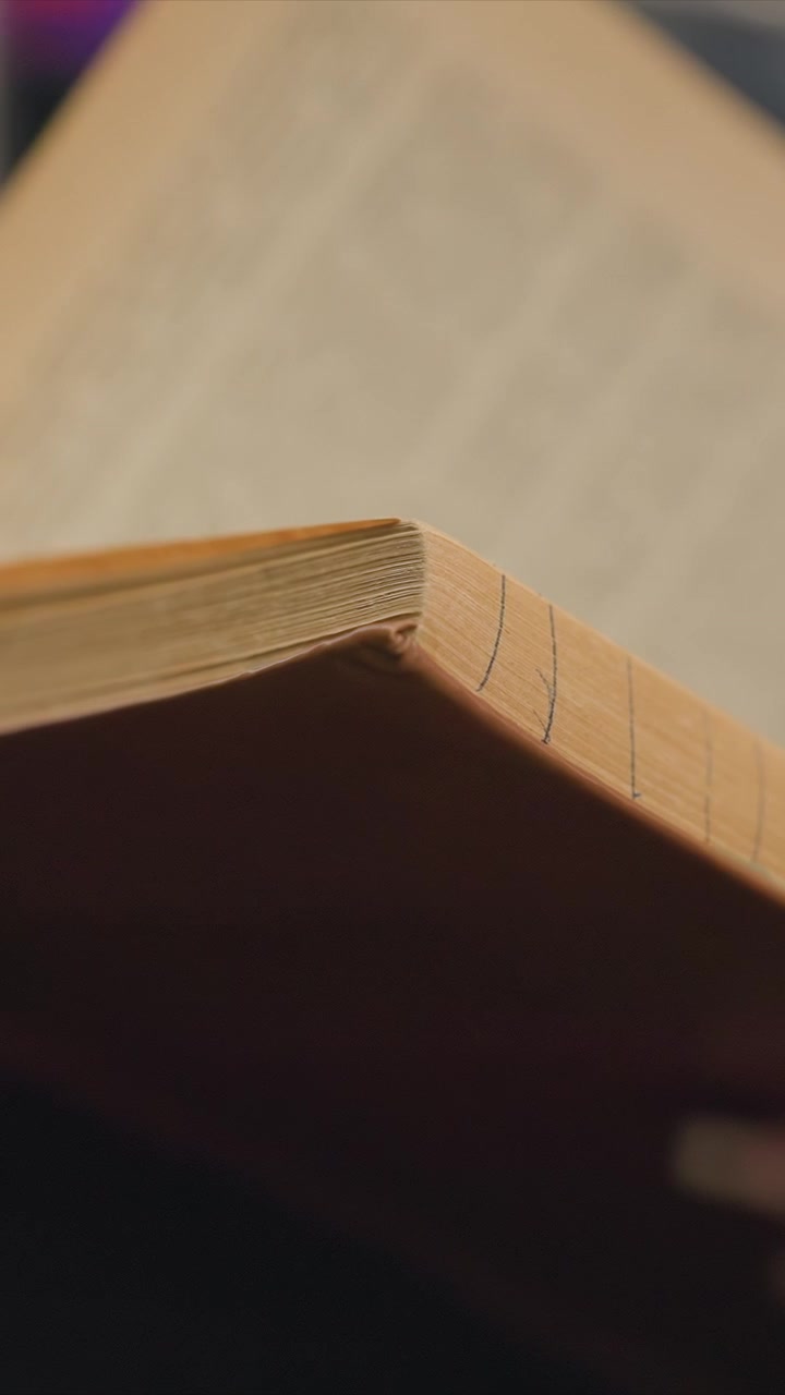 Close up of book corner highlighting its worn pages. Pages show faint blue lines suggesting annotations or markings. Scene is set indoors with blurred background elements