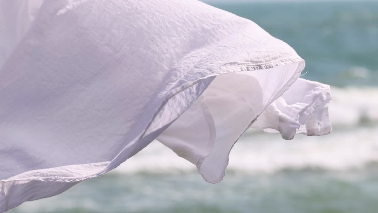 A white fabric gracefully moves with the breeze, set against rolling ocean waves.