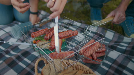 picnic at nature people are grilling sausages on grid taking fried wurst by hands and knife closeup view