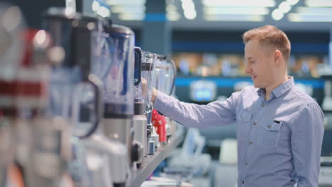 Young-handsome-man-in-appliances-store-chooses-a-blender-for-his-kitchen-looking-and-holding-various-models-in-his-hands