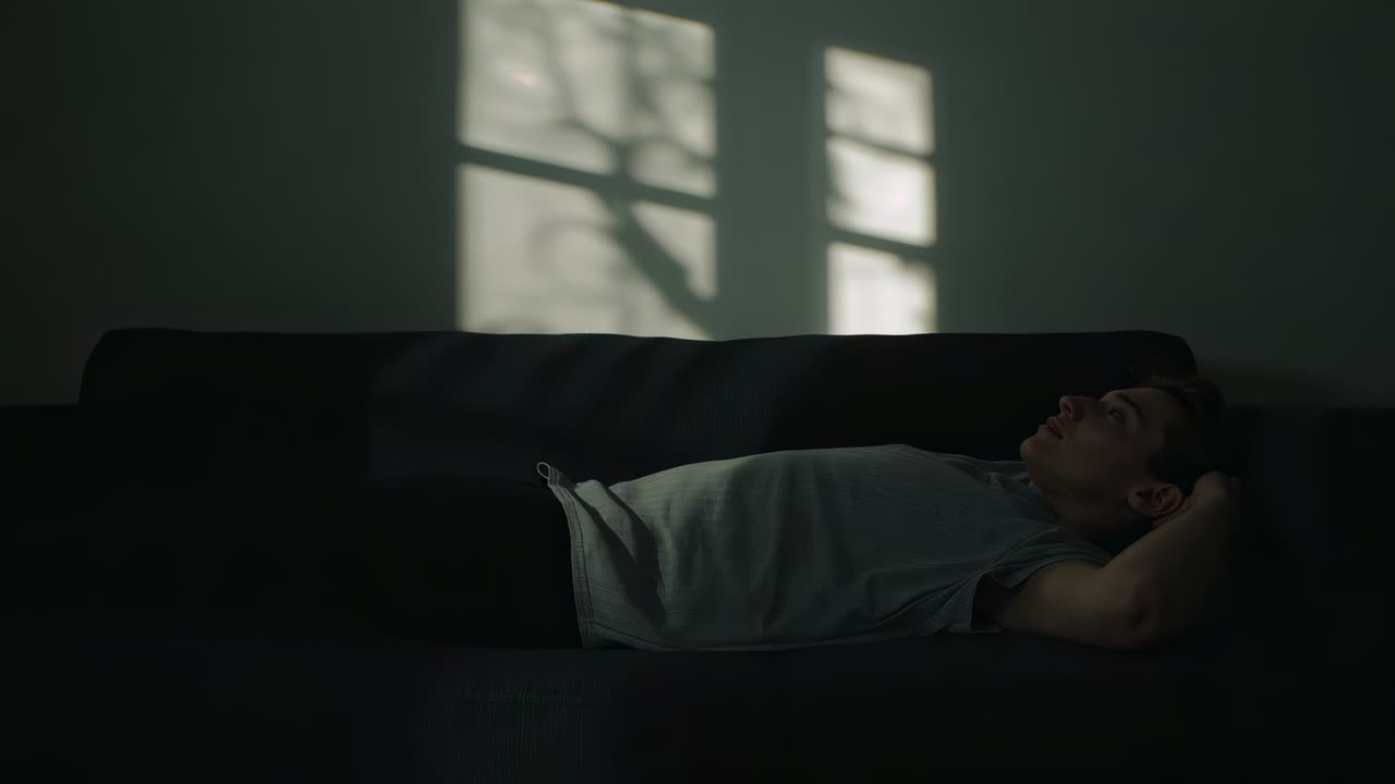 Lying man in light tee breathing on sofa at home, sunlight casting branch shadow, copy space