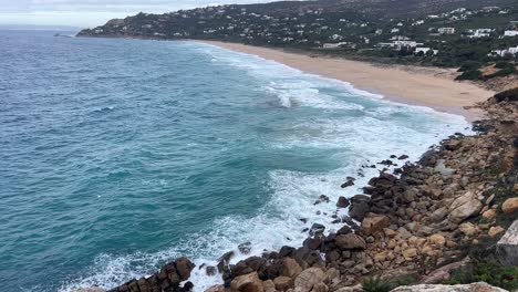 Slow-motion-of-coastal-sea-waters,-waves-are-crashing-at-shore-banks,-in-distance-visible-residential-area-with-houses