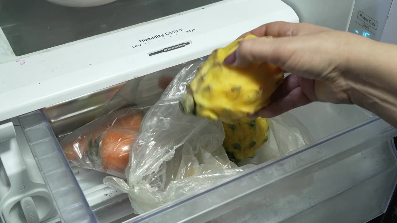 Eine Person greift in ein Kühlschrank-Gemüsefach und holt eine gelbe Drachenfrucht aus einer Plastiktüte neben gelagerten Karotten. Nützlich für Themen wie Lebensmittelaufbewahrung, Ernährung oder häuslicher Lebensstil.