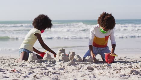 african american children wearing face masks playing with sand on the beach