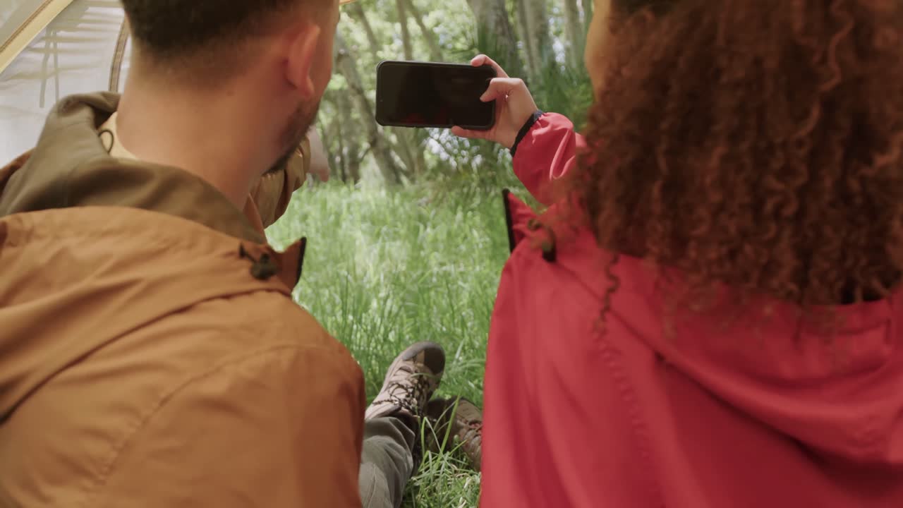 pareja afroamericana sentada en una tienda de campaña, usando un teléfono inteligente con espacio de copia en el bosque, cámara lenta