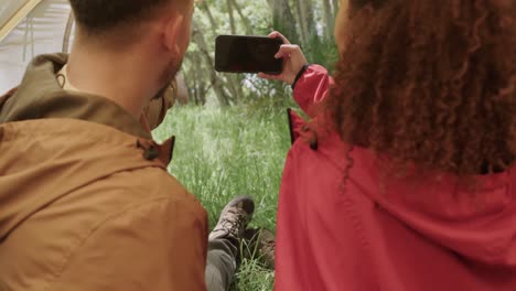 african american couple sitting in tent, using smartphone with copy space in forest, slow motion