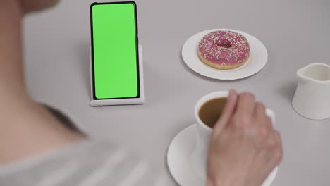 woman sitting at table coffee donut using smartphone with chroma key green screen, scrolling through social network media online shop internet. smartphone in horizontal mode with green screen mock-up.