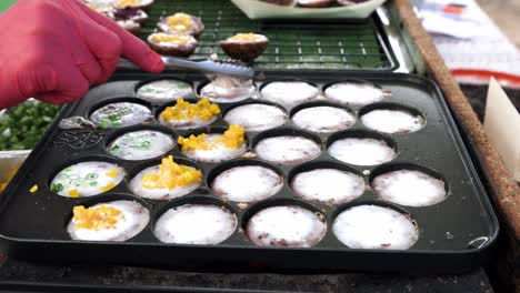 El-Vendedor-De-Comida-Callejera-Está-Preparando-Kanom-Krok-O-Pudín-De-Coco-Tailandés,-Una-Especie-De-Postre-merienda-Tailandés-Antiguo-Con-Opciones-De-Coberturas-Como-Cebolleta,-Granos-De-Maíz,-Taro,-Etc.