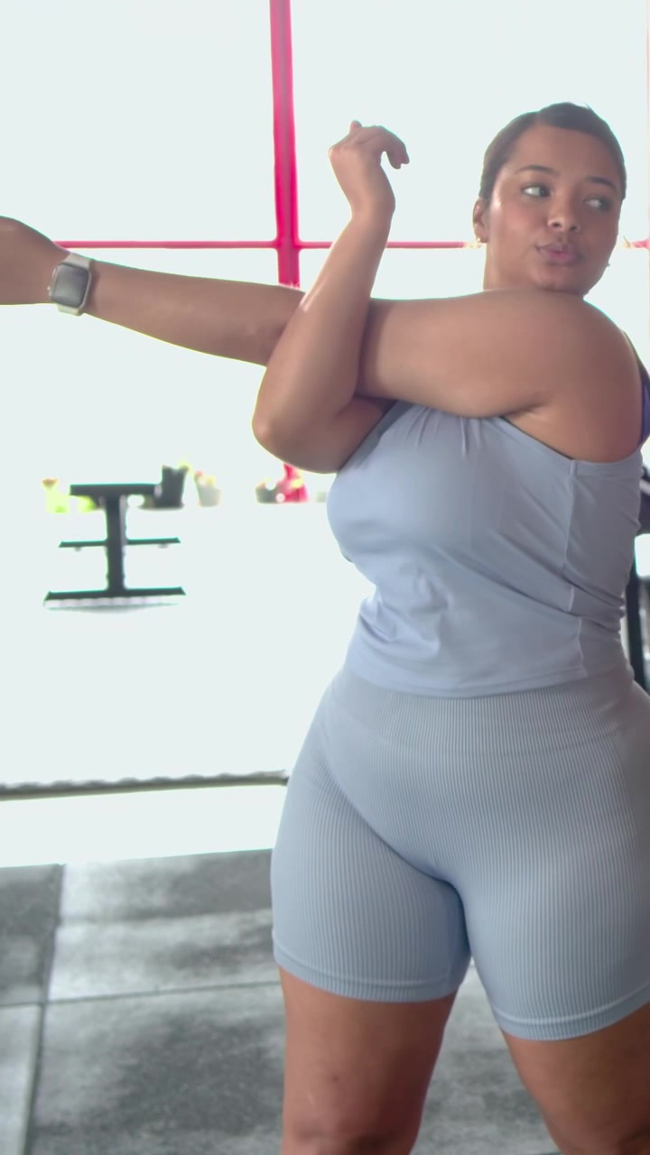 Video verticale: Inizio riscaldamento, donna in completo blu che esegue allungamenti a braccia incrociate in palestra, controllando l'orologio.