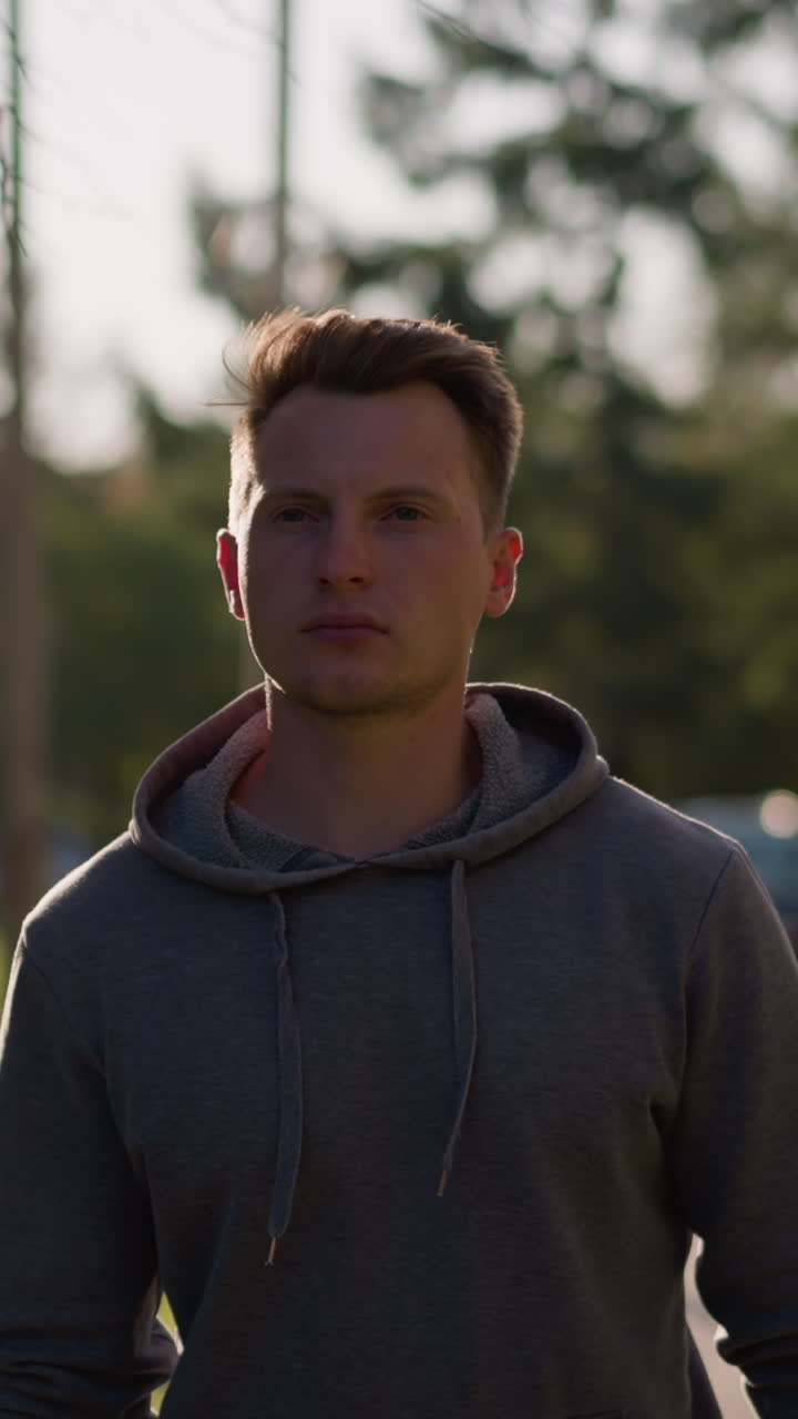 Young man in dark gray hoodie walking outdoors in sunny park with serious expression, surrounded by green grass, trees, distant building in soft focus background, autumn setting, relaxed mood