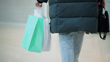 close-up back view of person in jeans trousers and black jacket walking in well-lit retail space carrying black handbag and two shopping bags, blurred background with modern mall interior