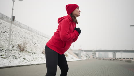 coach working out in winter, stretching into a focused side lunge on paved ground, arms clamped close to chest, wears hoodie and black leggings, exercising near snowy hill with urban surroundings