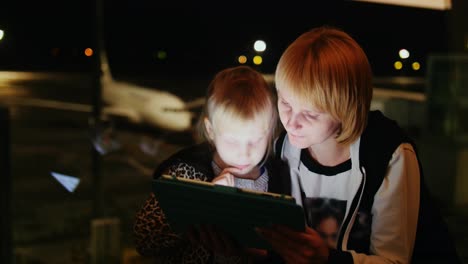 mamá e hija juegan en la tableta en el aeropuerto a última hora de la tarde, el avión es visible fuera del