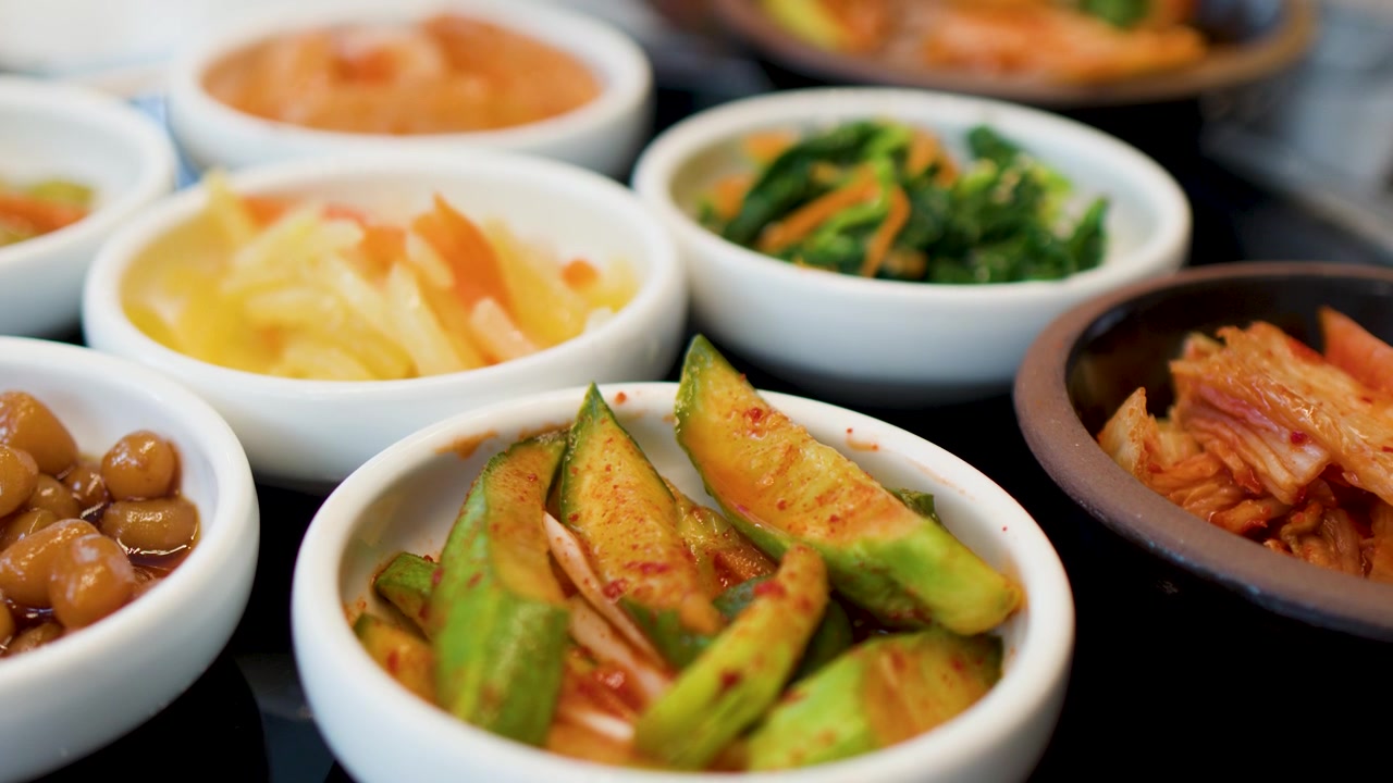Kamera close-up menyorot berbagai sayuran fermentasi, kimchi, dan hidangan pendamping berbumbu di atas meja.