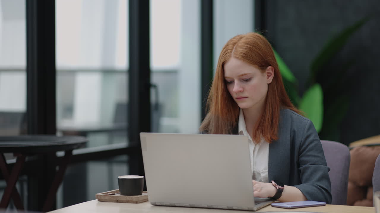 A young red-haired woman is working on a laptop. Remote work. Home office. Office work. A woman in a suit in the office is typing on a laptop