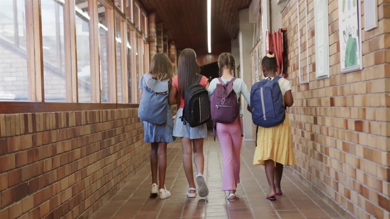vista trasera de alumnas felices y diversas con bolsas escolares caminando en el corredor de la escuela primaria