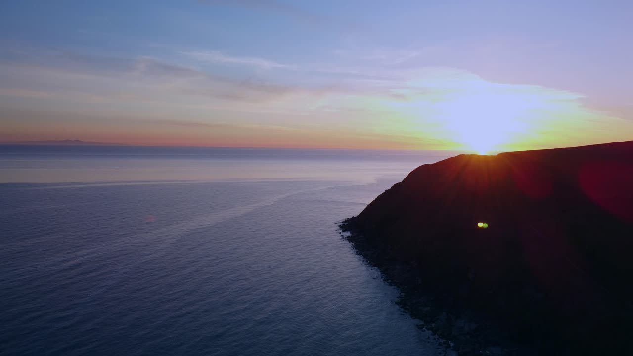 Serene aerial view of sun setting behind dark cliff over calm ocean waters.