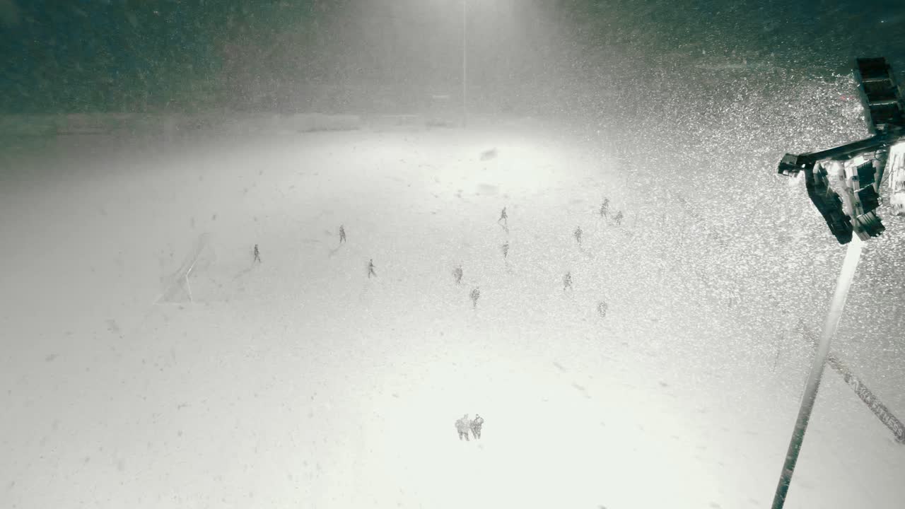 Aerial drone footage showing people playing soccer or football in a snowstorm or blizzard during night or evening time while a large and bright projector light lights up the soccer field. Snowflakes.