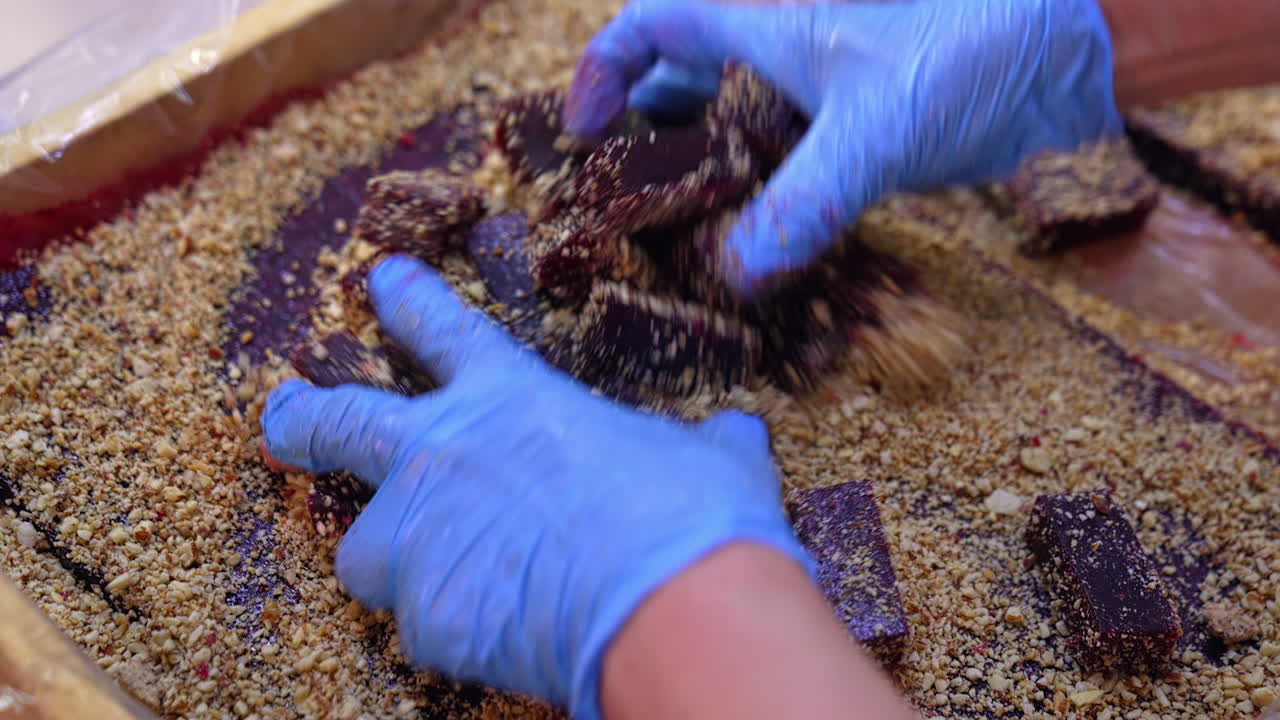 Hands in blue gloves cover little bars of marmalade in nuts. Purple jelly bars being mixed up with chopped walnut. Side close up.