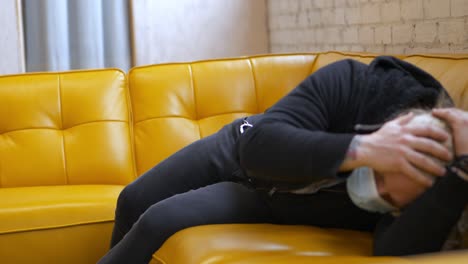masked long blonde hair bearded man falling passing out on yellow vegan leather vinyl couch feeling sick dying ill then gets up when called on and limps away - in cinema 4k (60fps real time).